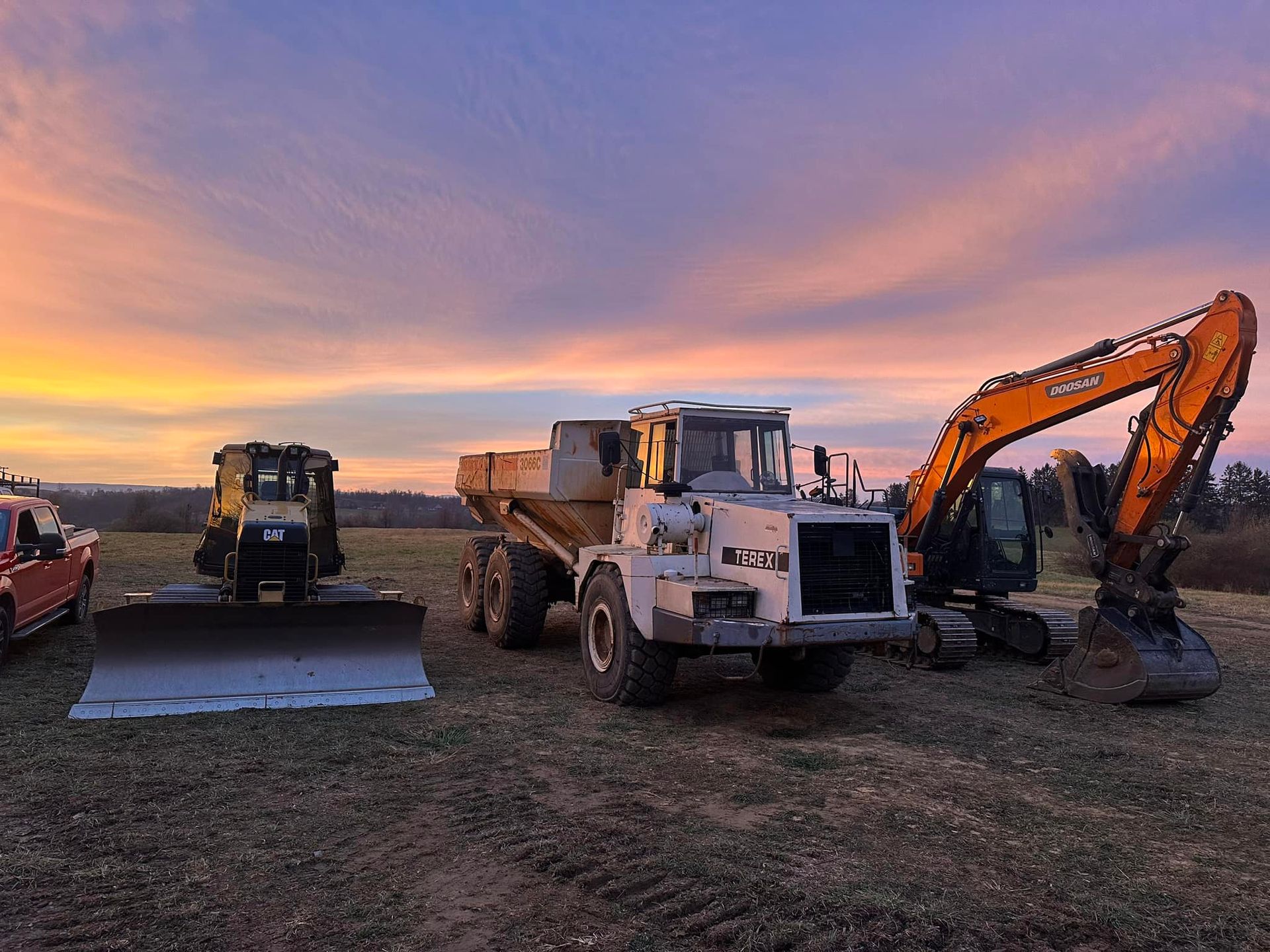 Heavy construction equipment including a bulldozer, dump truck, and excavator parked in a field at sunset.