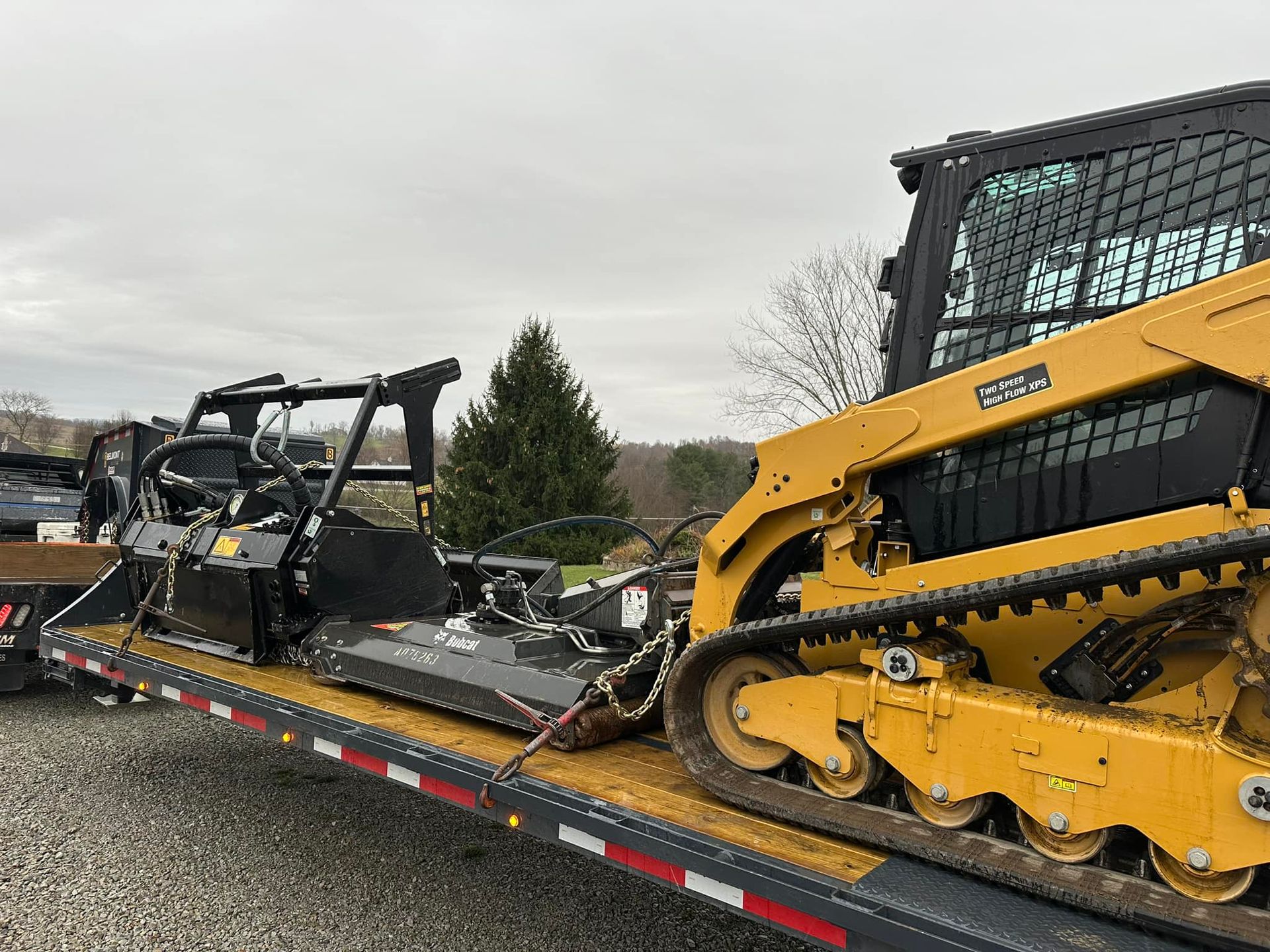 A yellow tracked skid steer loader sits on a flatbed trailer next to a black industrial brush cutter attachment.