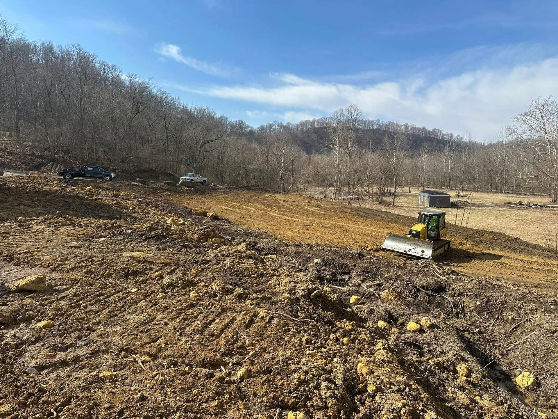 A yellow bulldozer sits on a freshly cleared dirt lot, with trucks nearby and a forest-covered hillside in the background.