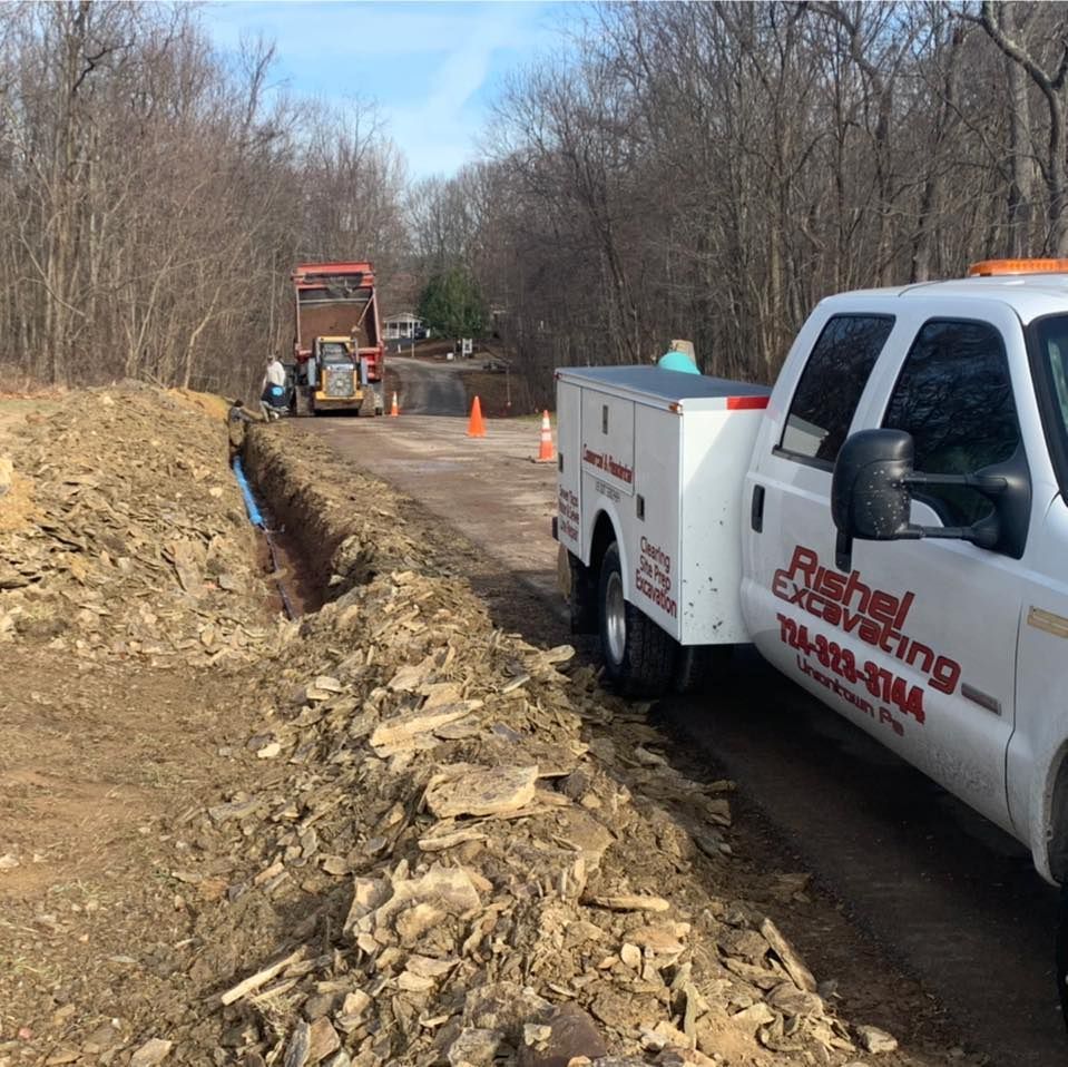 A Rishel Excavating truck parked beside a trench under construction on a dirt road, with heavy machinery in the distance.