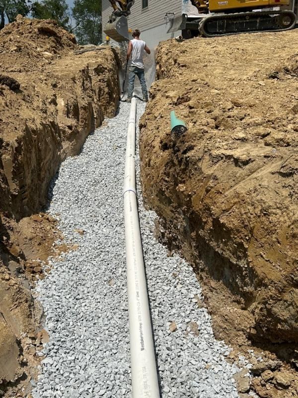 A person stands in a deep trench lined with gravel, where a white pipe is being installed near construction equipment.