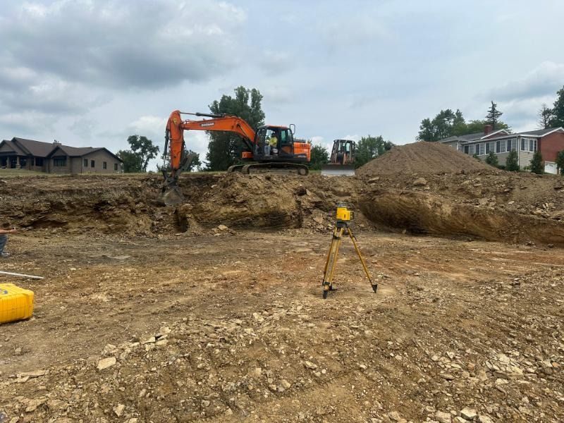 An orange excavator works at a construction site with a tripod survey tool in the foreground under a cloudy sky.