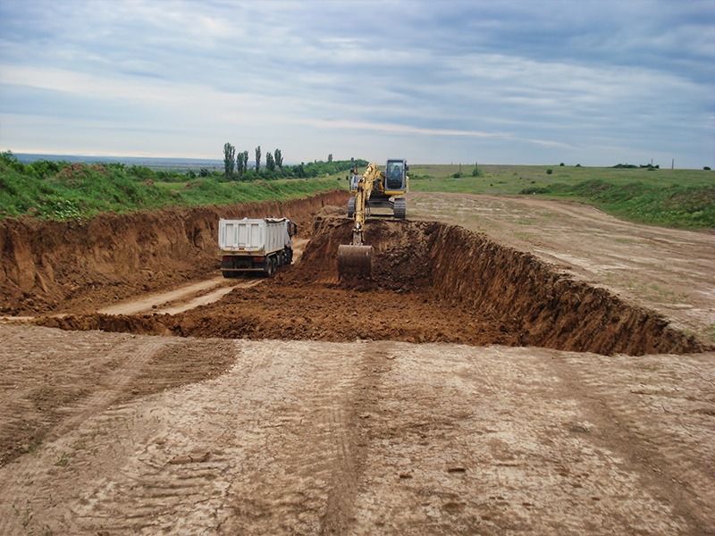 An excavator digs a trench in an open field, loading soil into a waiting dump truck.