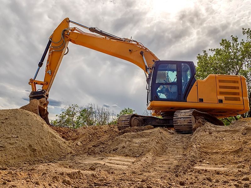 A yellow excavator rests on top of a dirt pile at a construction site against a cloudy sky.