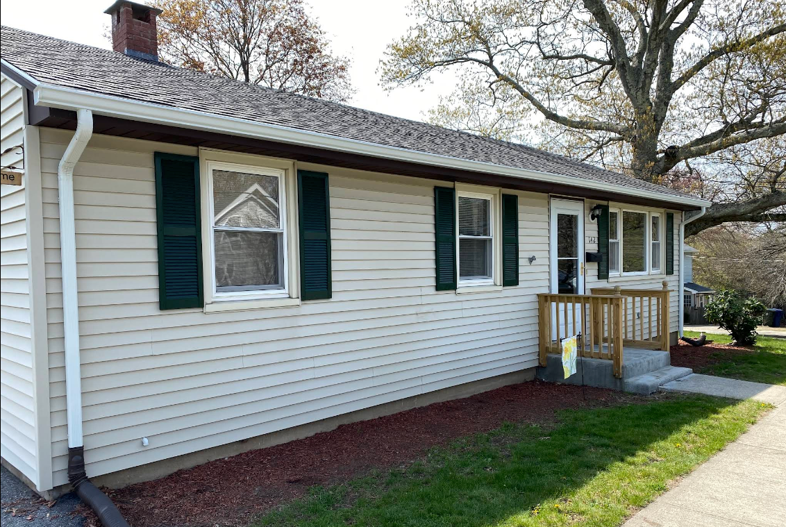Single-story white house with green shutters, front porch, and brick foundation in a sunny yard