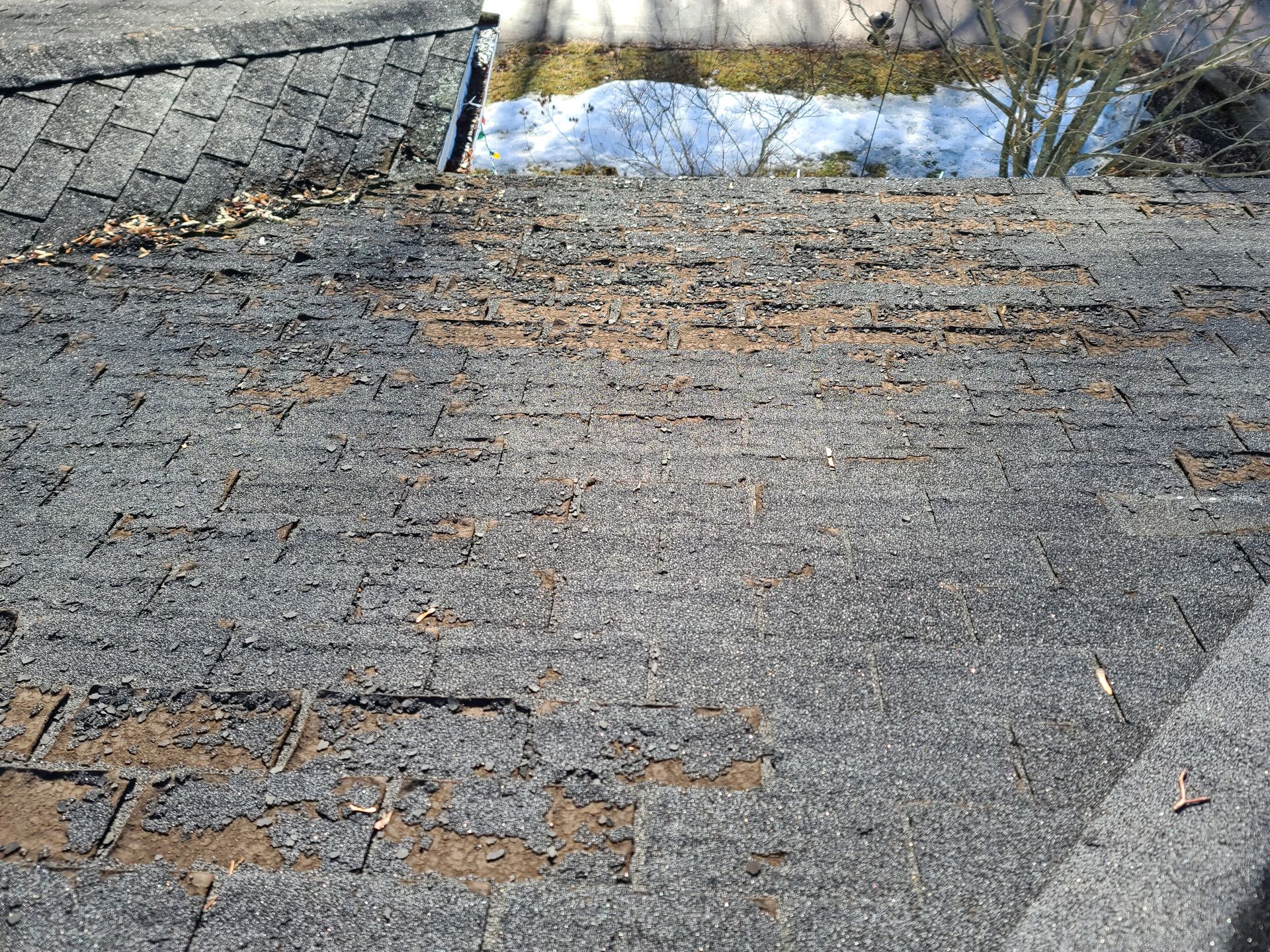 Damaged gray shingled roof with missing patches and debris near a gutter opening