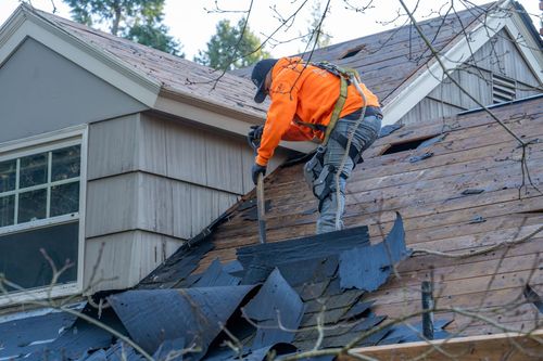 Worker in orange jacket repairs a house roof with a hammer next to a skylight