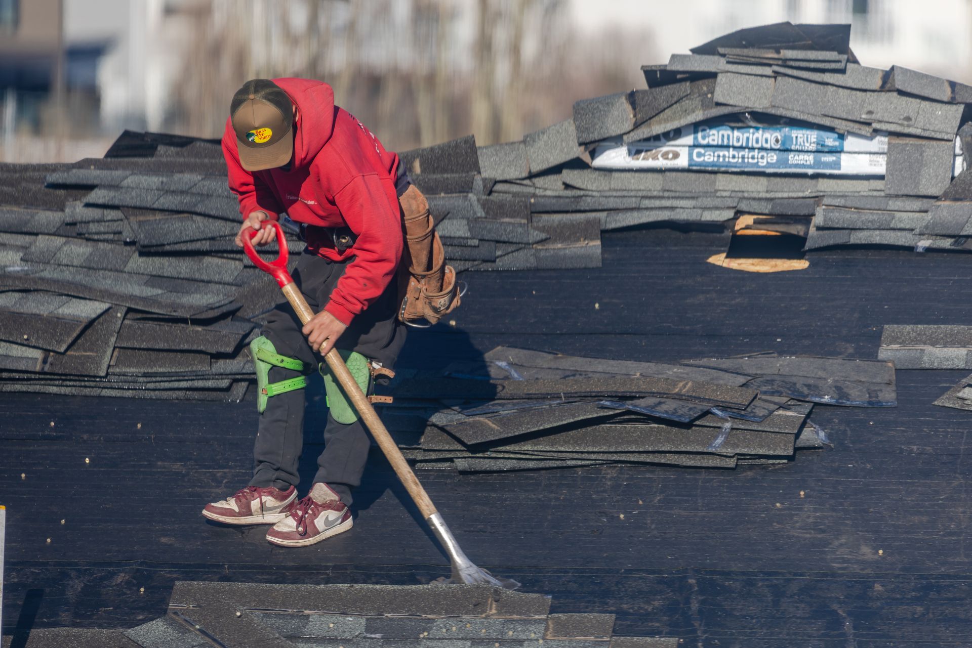 Worker in a red hoodie shoveling debris on a rooftop with stacked shingles nearby