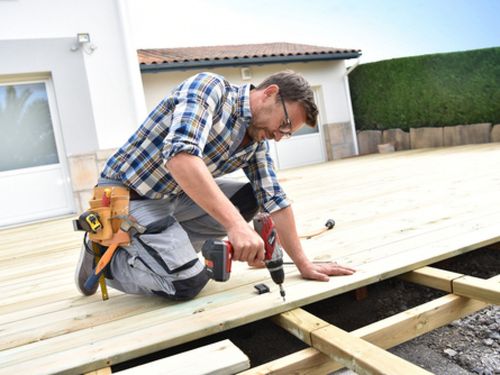 Worker kneeling on a wooden deck, using a drill to fasten boards outdoors.