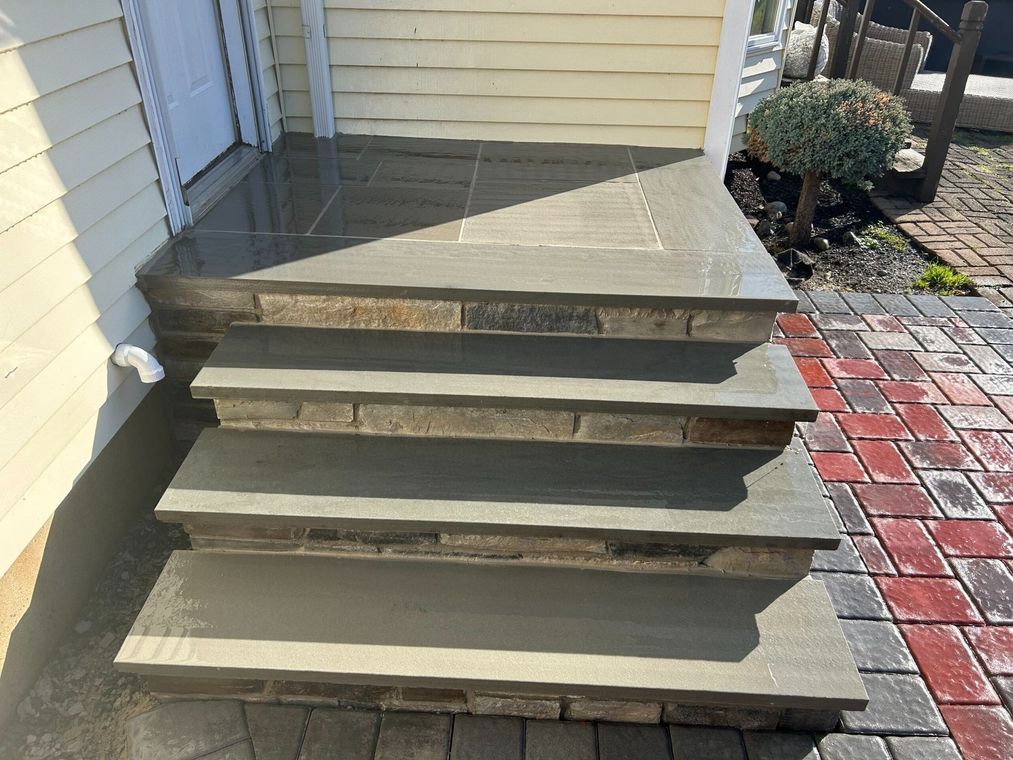 Stone steps leading up to a doorway, flanked by brick paving, with a small bush nearby.