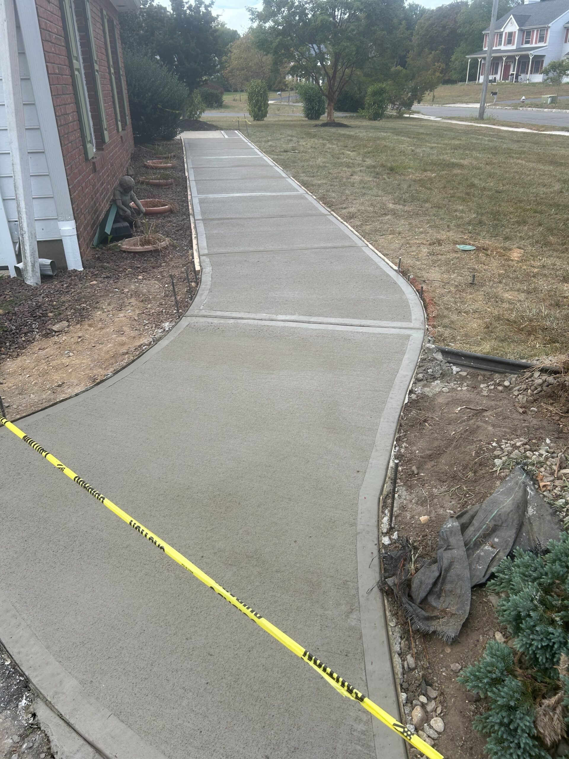 Newly poured concrete sidewalk with yellow caution tape. Next to a brick house and green grass.
