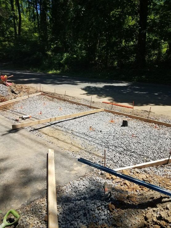 Construction site with gravel-filled formwork for a concrete slab, next to a paved road in a wooded area.