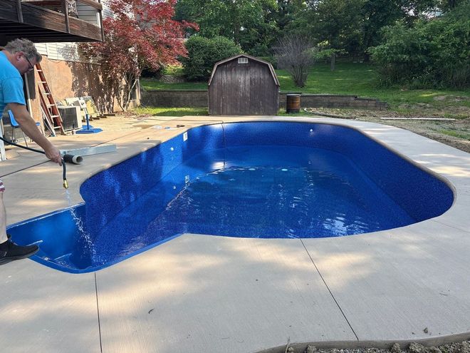 Person pressure-washes a backyard pool deck beside a blue in-ground pool.