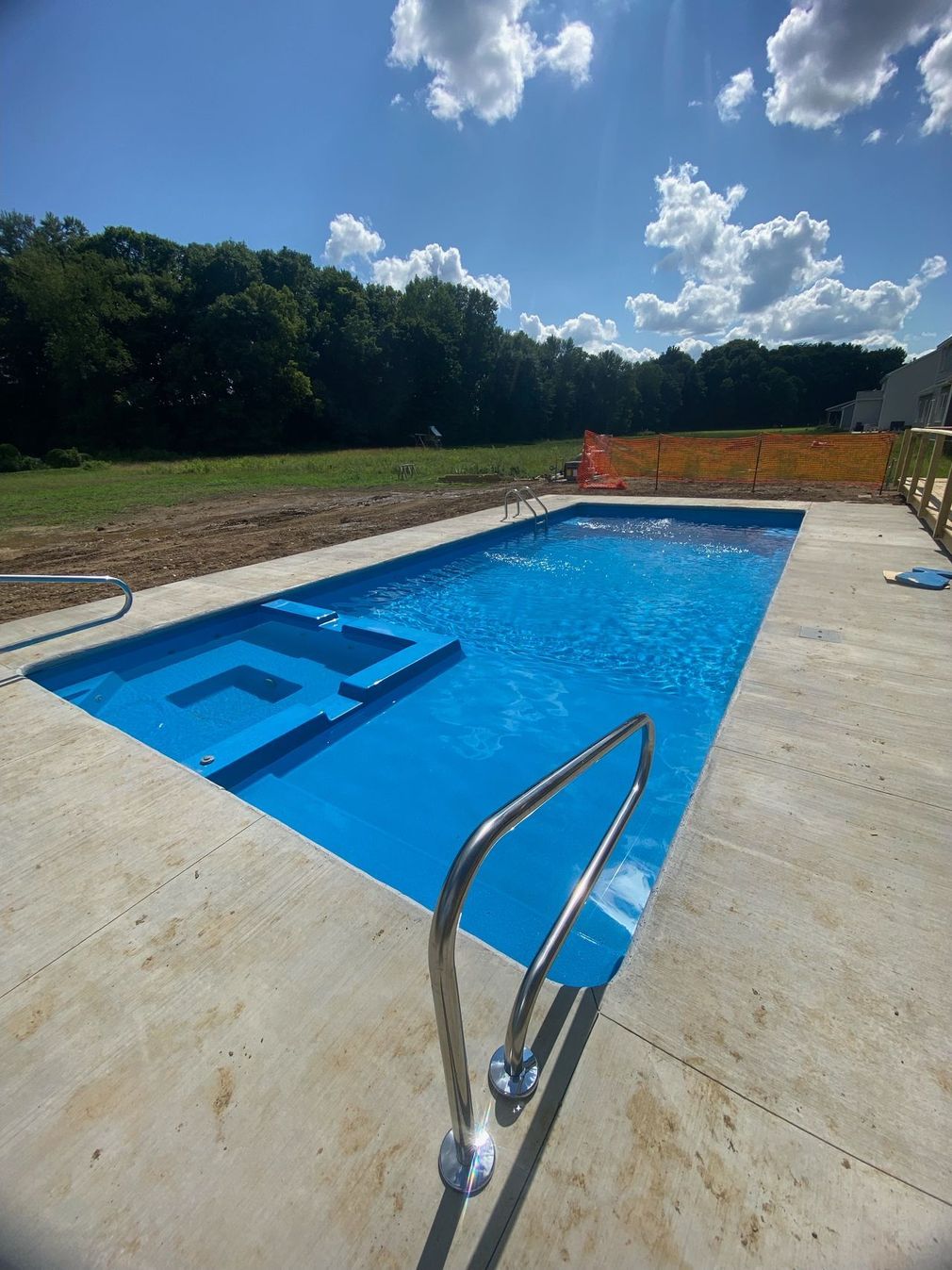 Backyard pool with steps and metal handrail under a blue sky, surrounded by concrete deck and trees