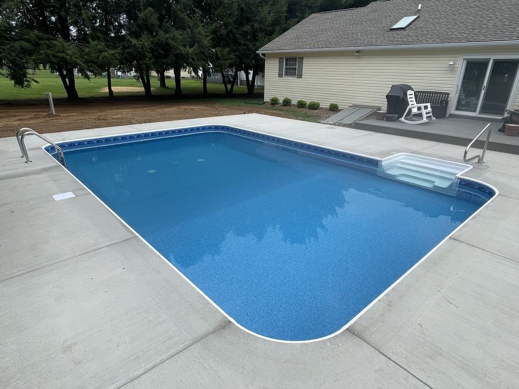Backyard in-ground pool with blue cover, concrete deck, and house patio chairs nearby