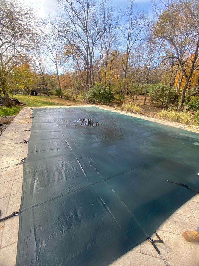 Covered backyard pool with black tarp, surrounded by autumn trees and a stone patio.
