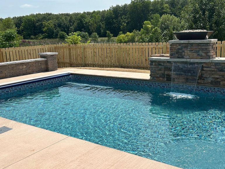 Backyard swimming pool with blue water, stone patio, and fenced yard with trees beyond.