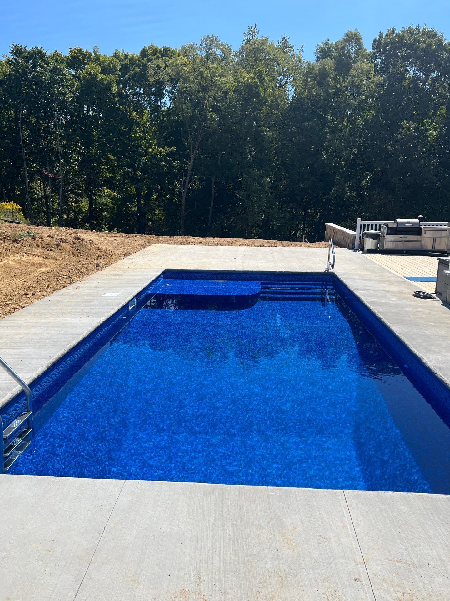 Rectangular blue swimming pool with a white deck and trees in the background