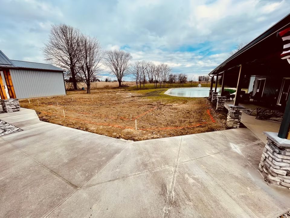 Wide concrete patio beside a barn and shed, opening onto a grassy yard with leafless trees and a small pond.