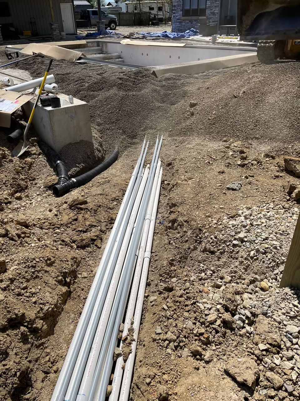 Construction site with gravel trench, long white pipes, and concrete footing in the background