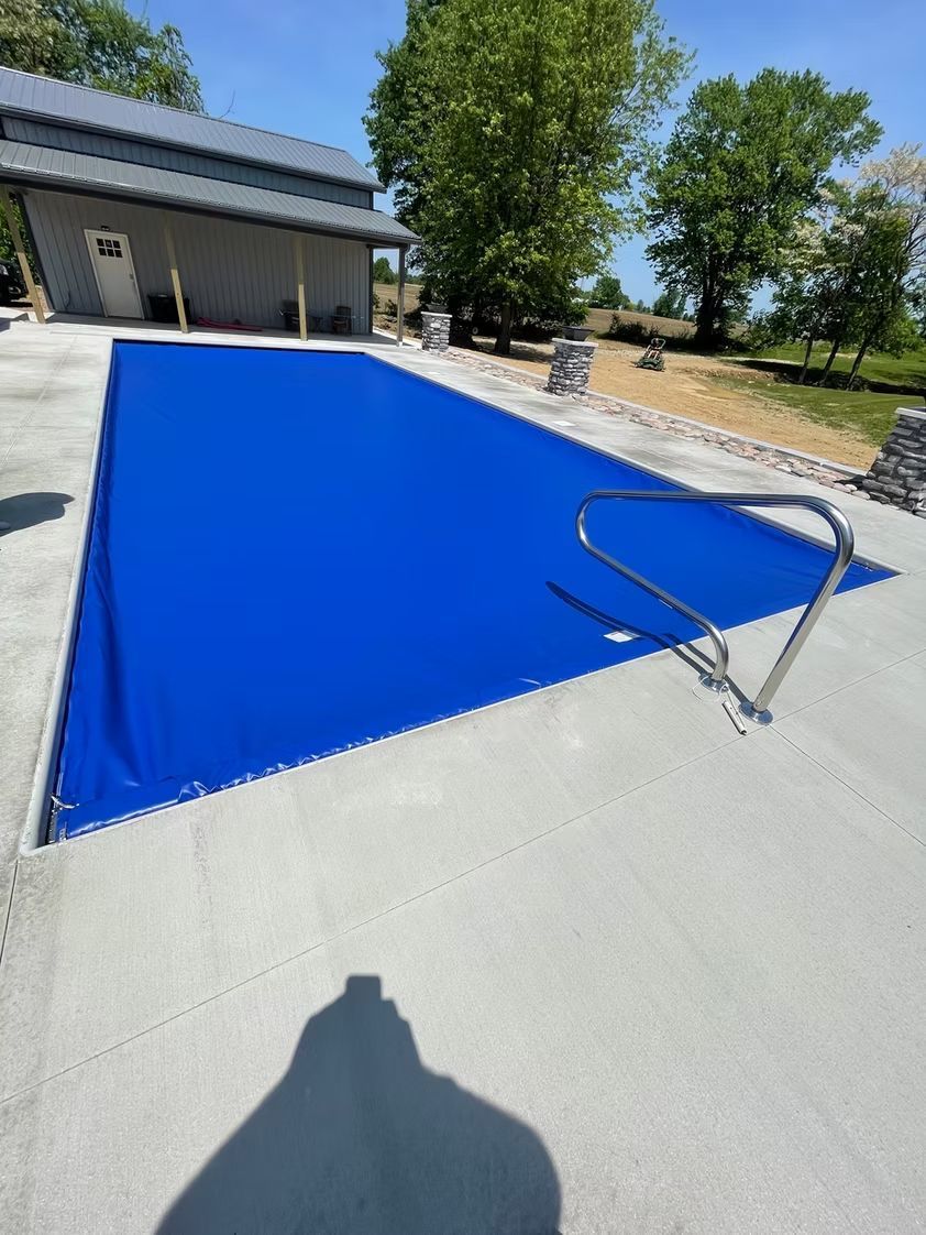 Blue pool cover stretched over a backyard swimming pool beside a concrete patio and house garagem