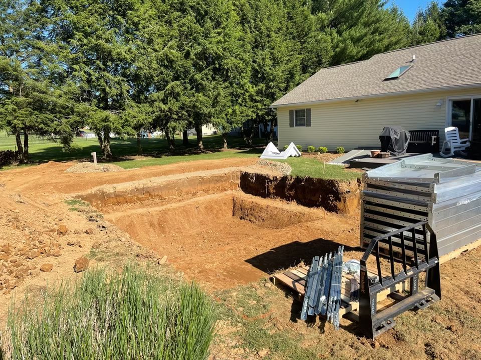 Backyard excavation beside a house, with a deep dirt pit and stacked patio materials by the deck.