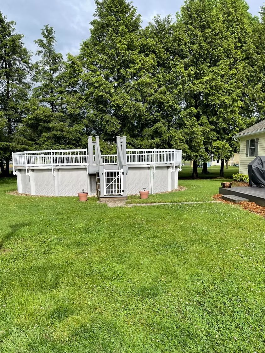 Above-ground pool in a grassy backyard with deck railing, trees, and a house beside it