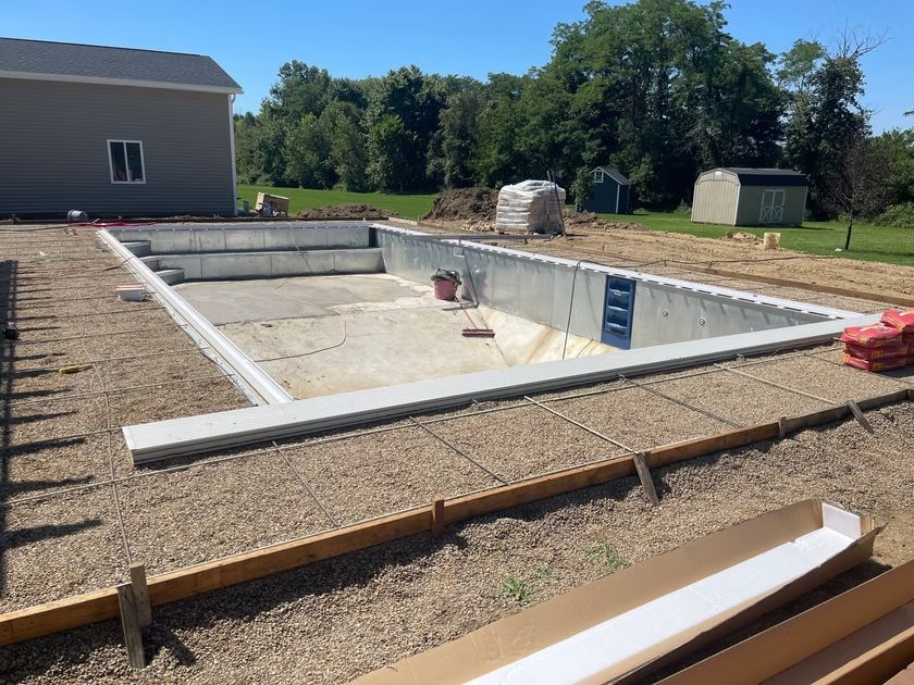 Rectangular in-ground pool under construction, with concrete walls and gravel yard near a house.