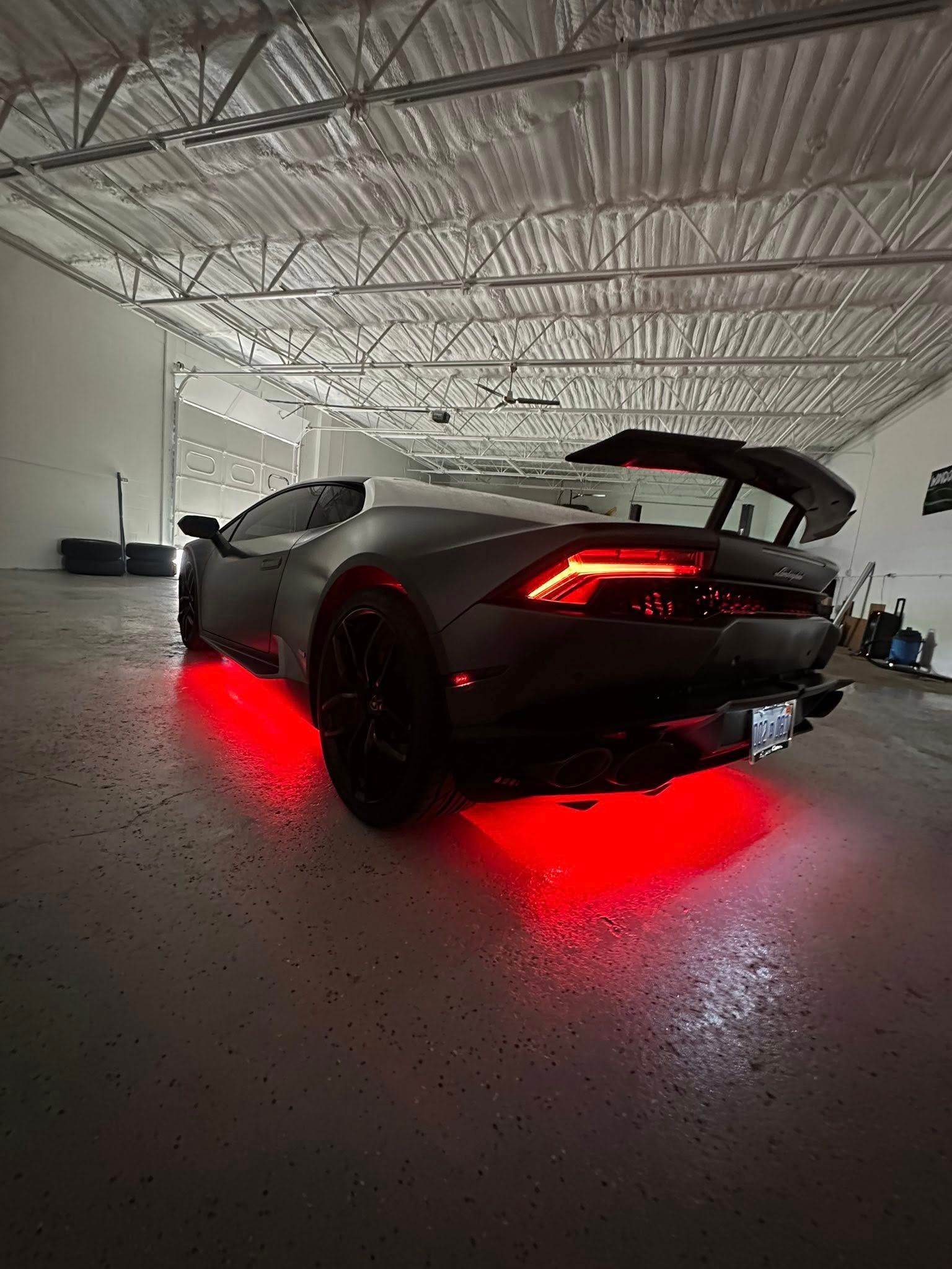 A dark grey sports car with a large rear wing and glowing red underglow lights parked inside a warehouse.