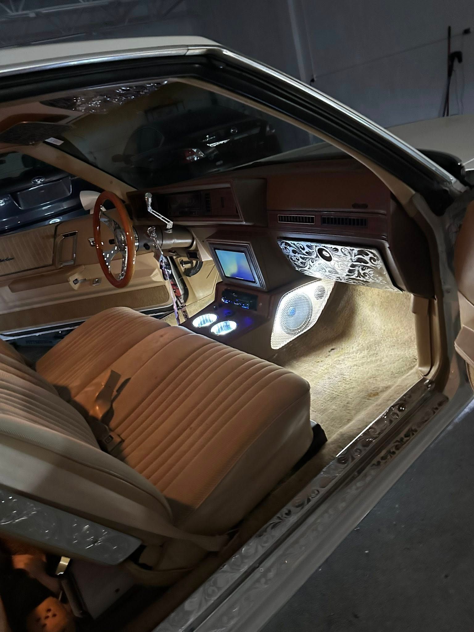 The tan interior of a modified car featuring a custom dashboard, light-up speakers, a wood-rimmed wheel, and ambient lights.