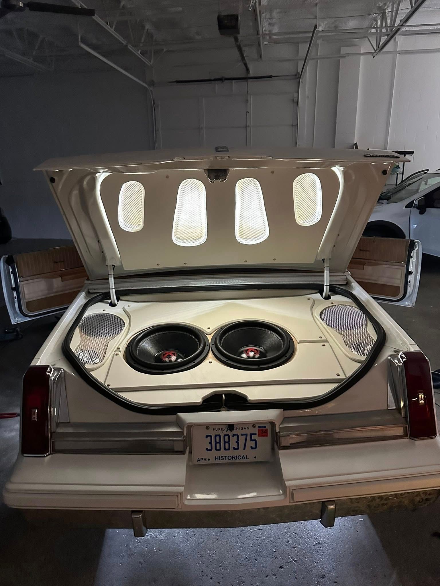 A custom white trunk interior of a car, featuring two large subwoofers, ambient lighting, and decorative speaker grilles.