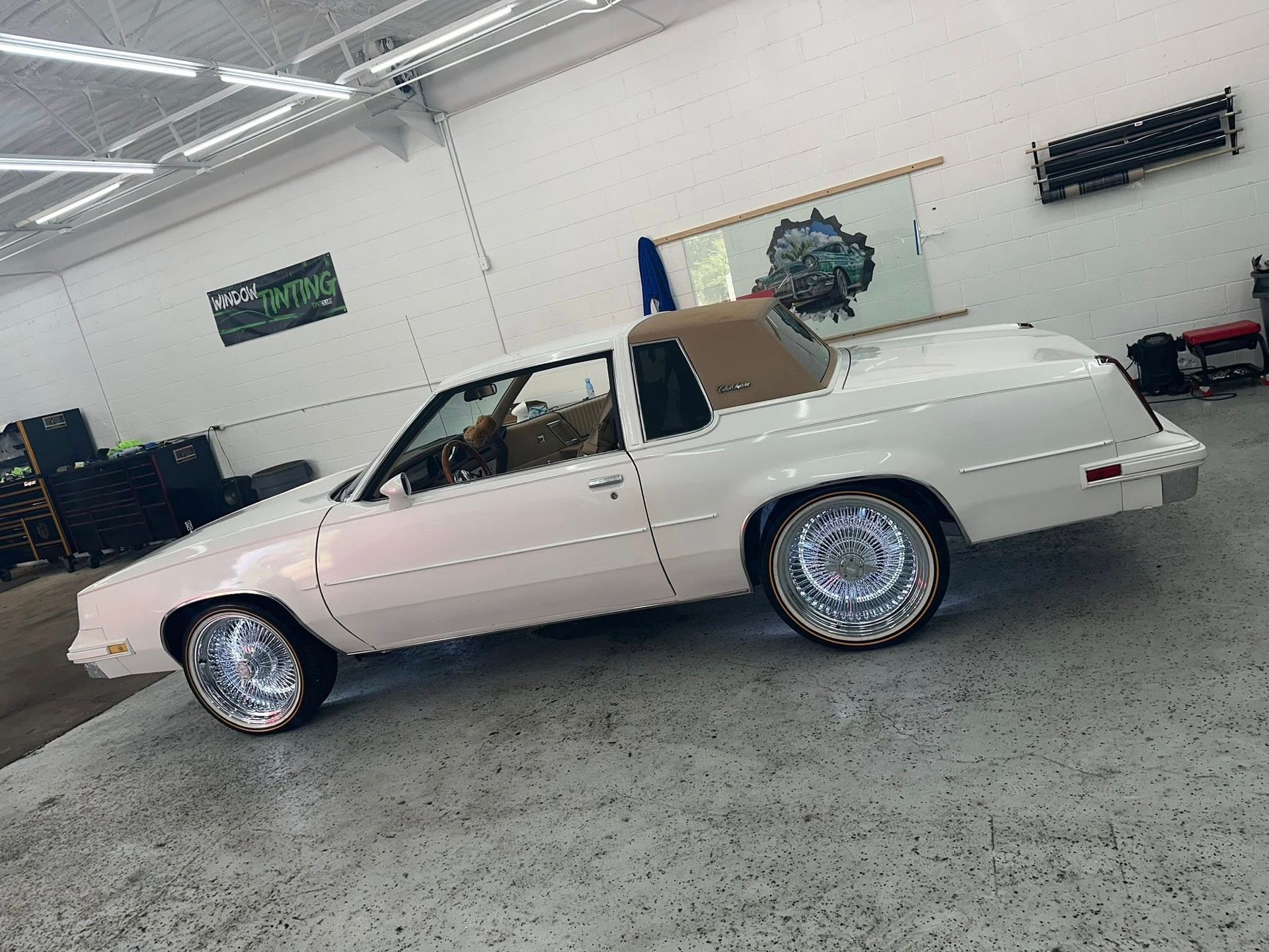 A cream-colored, vintage two-door coupe with a tan vinyl top and custom oversized chrome wheels, parked in a garage.