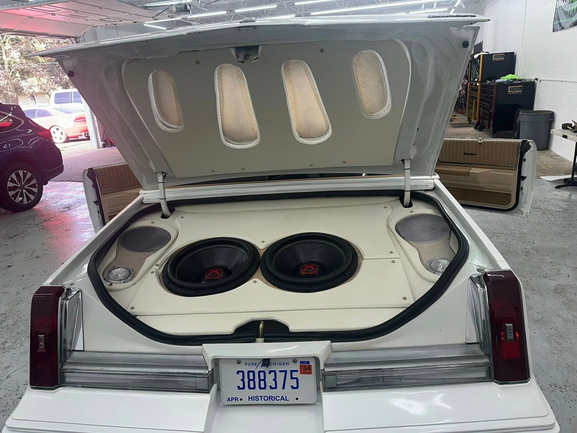 The open trunk of a white car reveals a custom audio setup featuring two large subwoofers and two speakers.