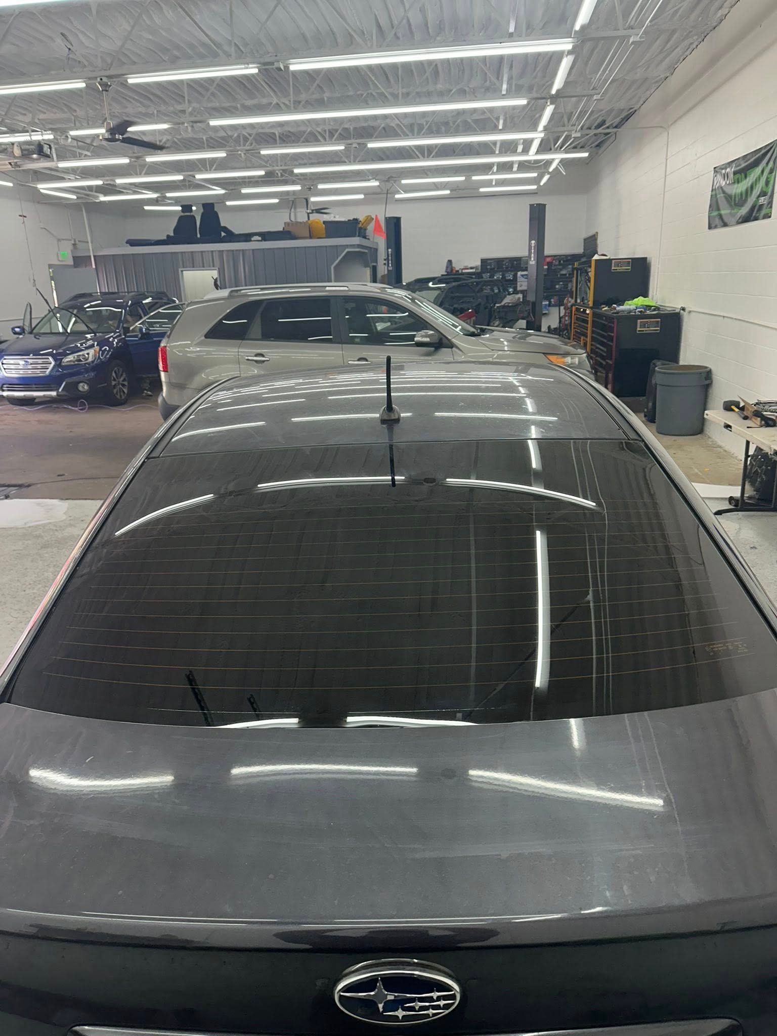 A dark gray Subaru parked in an auto repair shop with an overhead view of its tinted rear window and antenna.