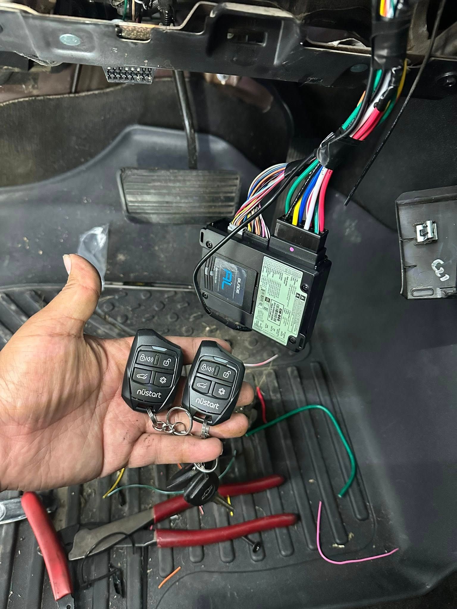 A hand holds two black remote car starters next to an open vehicle dashboard with exposed wiring and an electronic module.