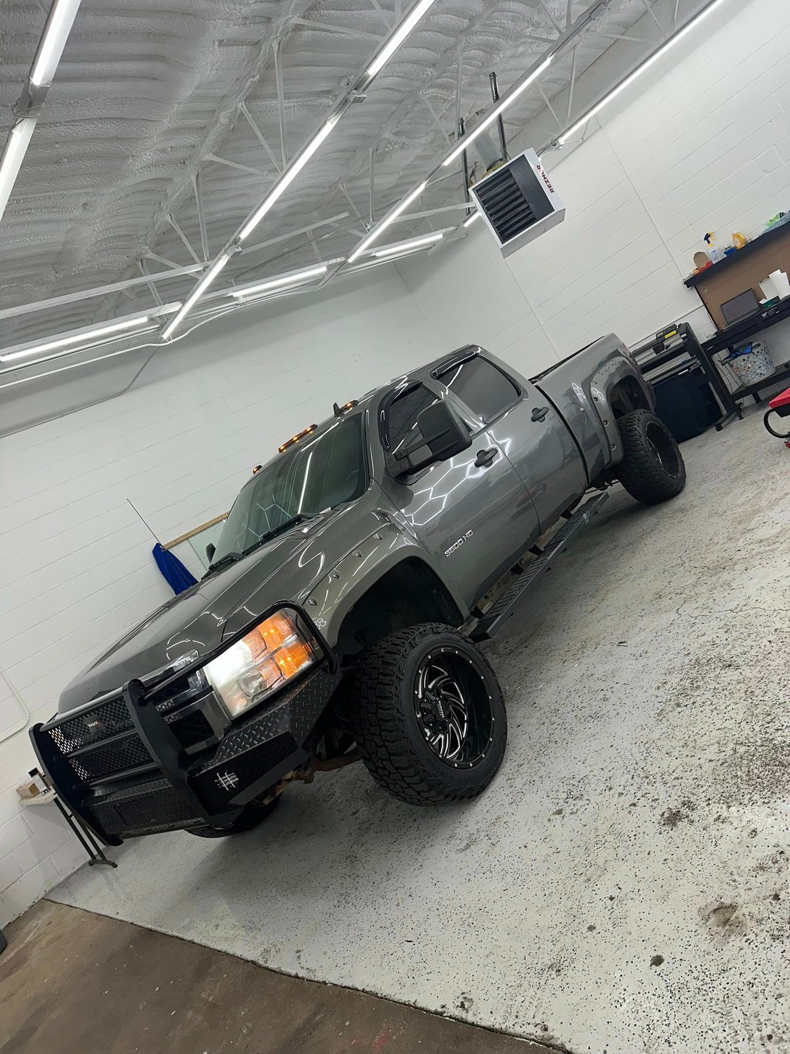 A gray lifted pickup truck with a black brush guard and large off-road tires, parked in a well-lit garage.