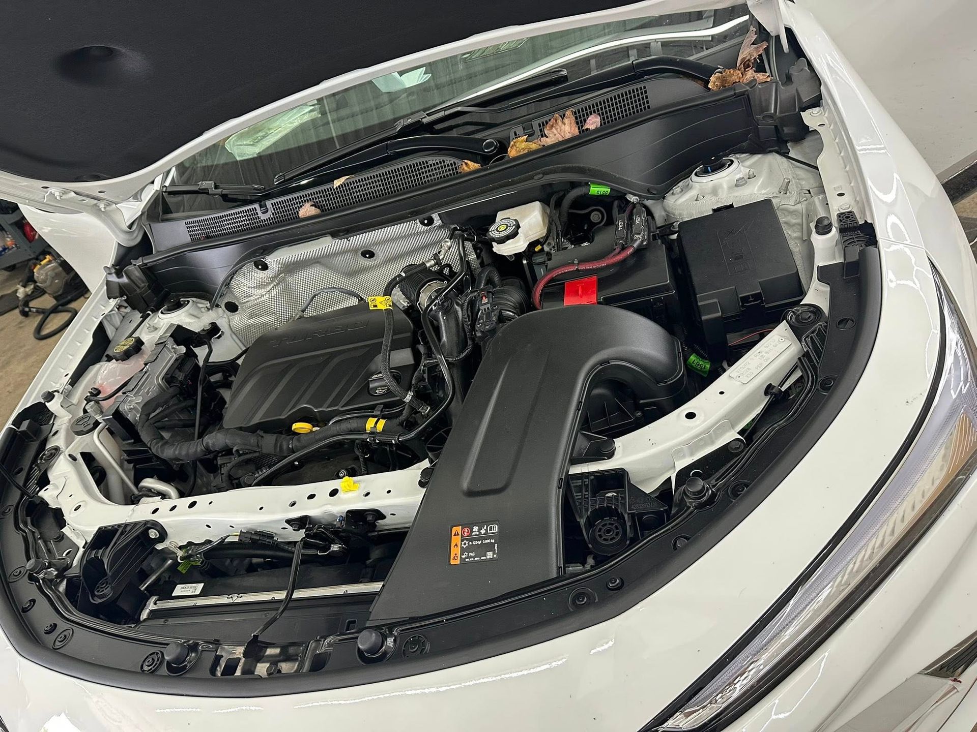 A high-angle view of an open white car hood revealing the engine bay, components, and a black air intake duct.