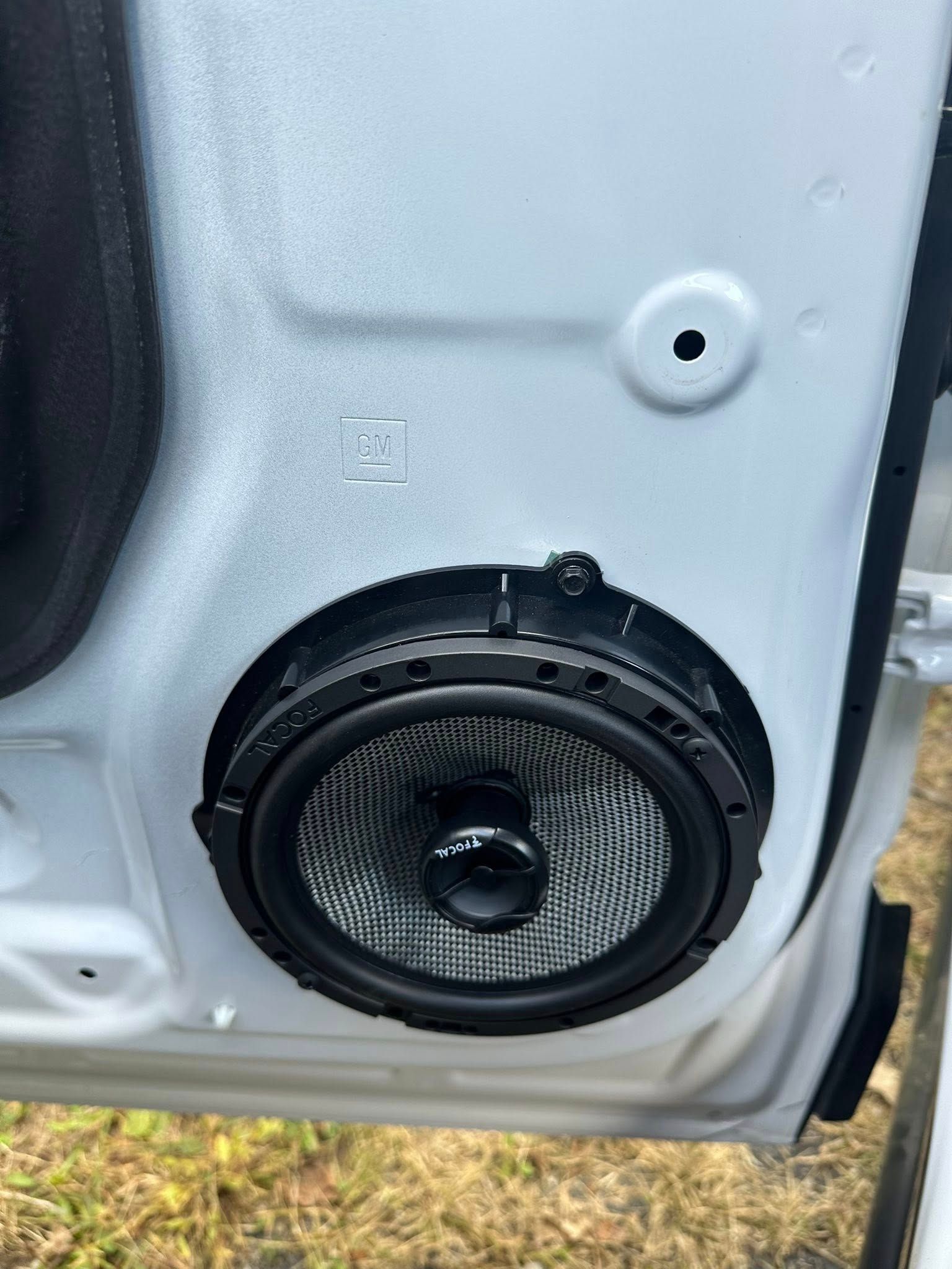 A circular car speaker with a black mesh cone mounted onto the white metal interior panel of a car door.