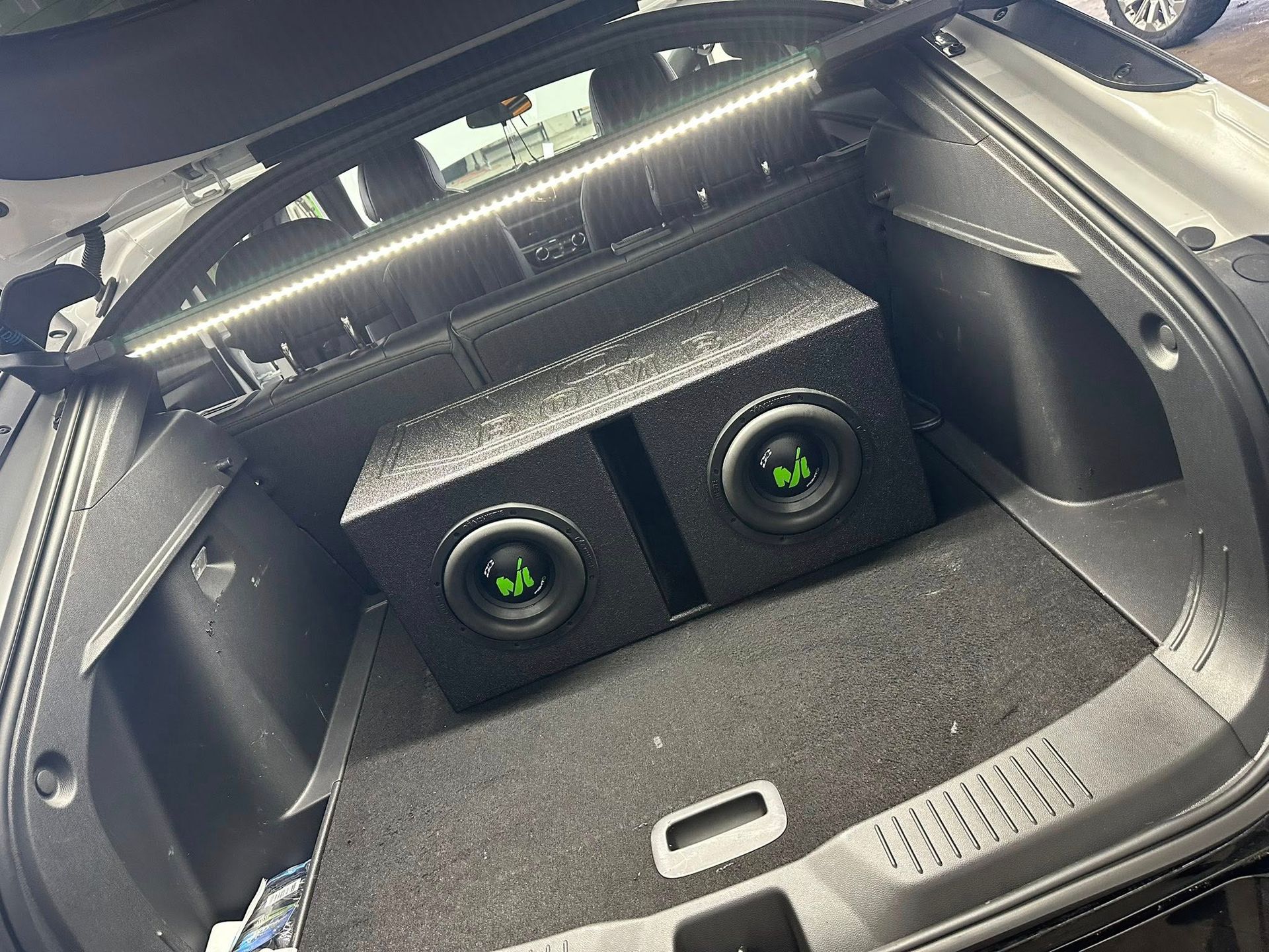 A car trunk interior featuring a dual-subwoofer speaker box with green logos and an overhead LED light strip.