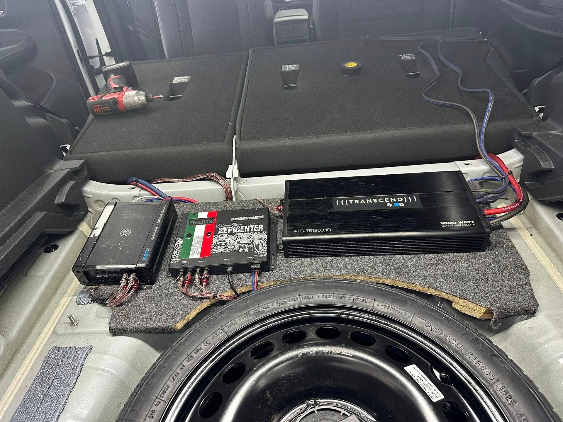 Car audio amplifiers and a distribution block mounted on a carpeted panel in a vehicle trunk above a spare tire.