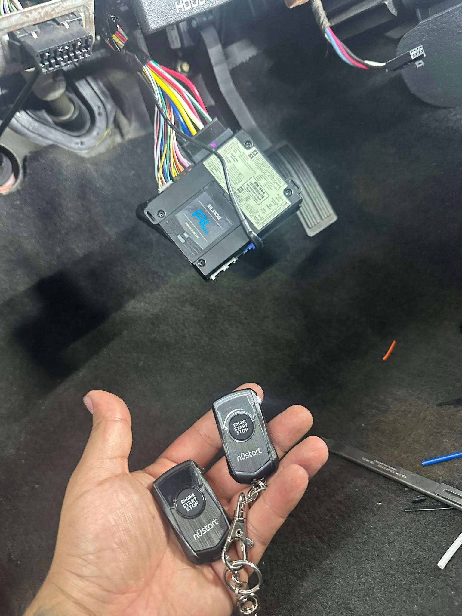 A hand holds two black automotive key fobs near a car's wiring harness and an electronic control unit under the dash.