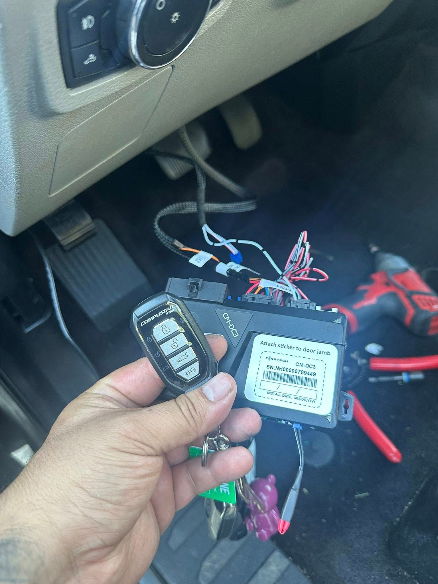A hand holds an electronic module with a keypad remote under a vehicle dashboard, with tools and wiring visible nearby.