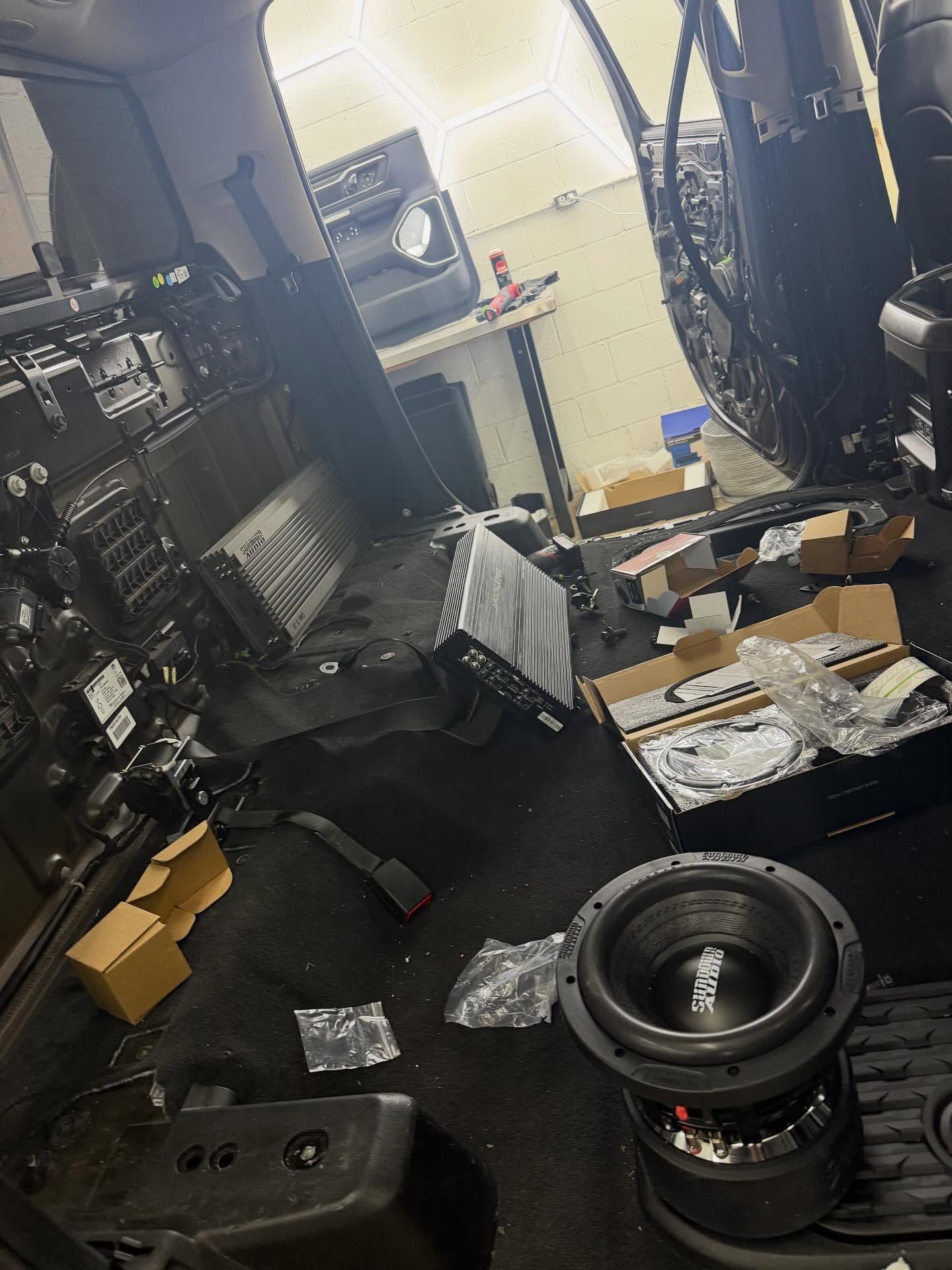 A car interior undergoing an audio system installation with a subwoofer, amplifier, and tools scattered on the floor.