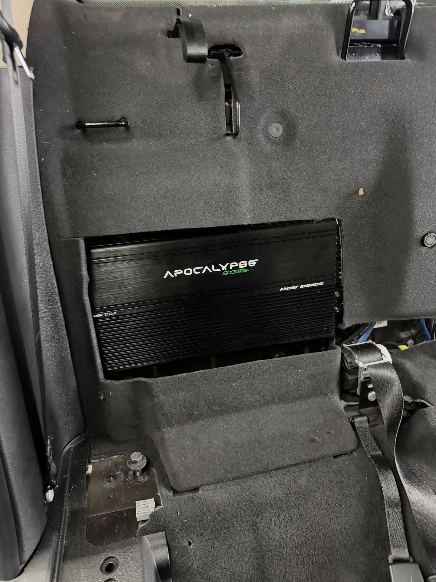 An Apocalypse brand car audio amplifier mounted in a black-carpeted interior panel behind a vehicle seat.