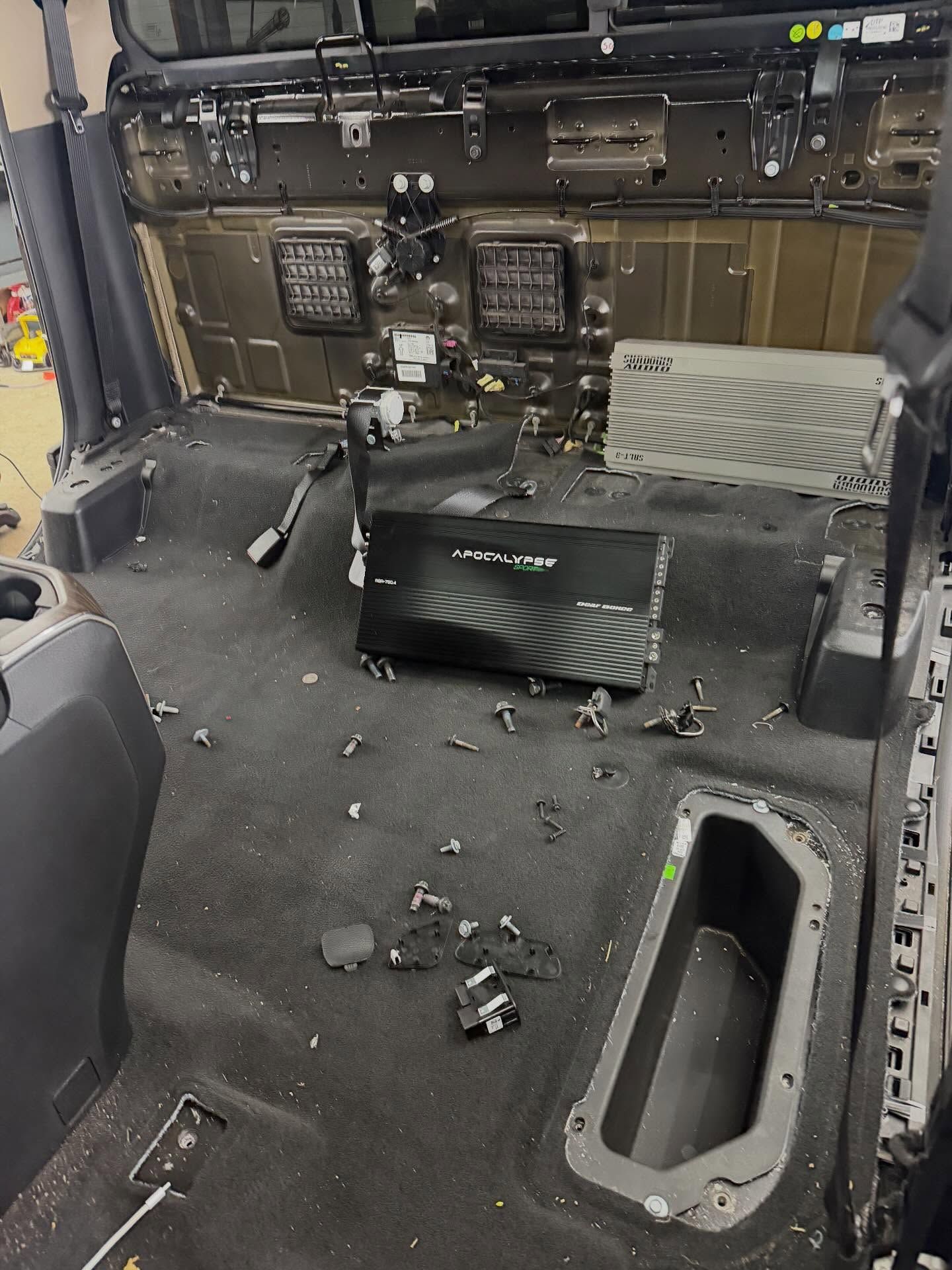 Interior view of a vehicle cab with seats removed, revealing two black audio amplifiers and scattered hardware on floor.