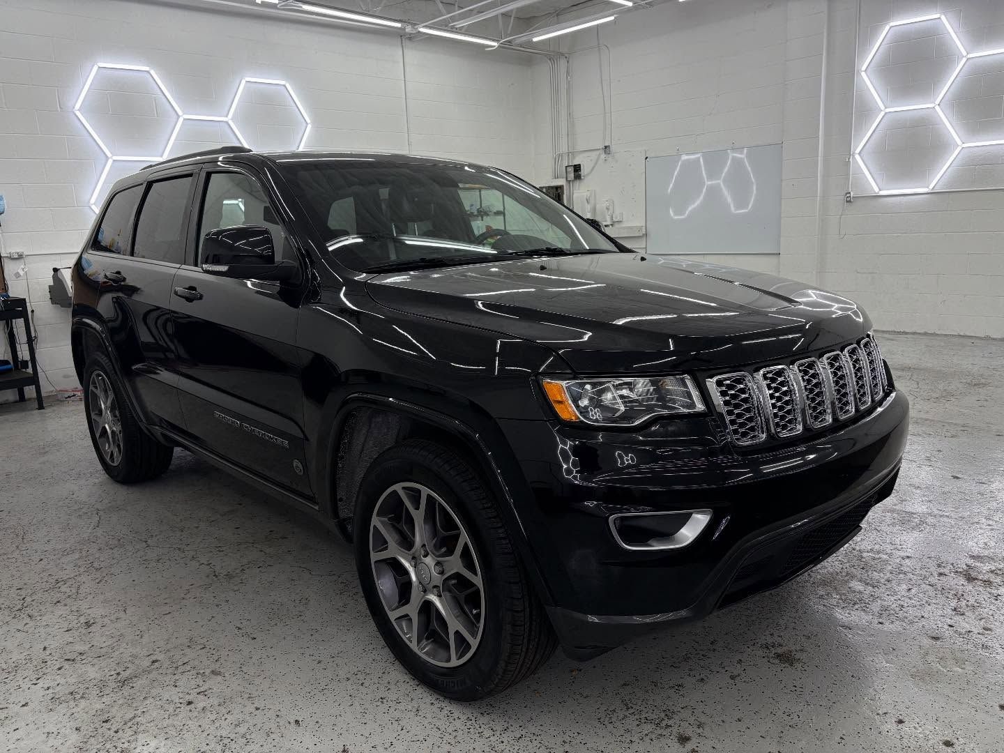 A black Jeep Grand Cherokee parked in a brightly lit professional garage with hexagonal wall lights.