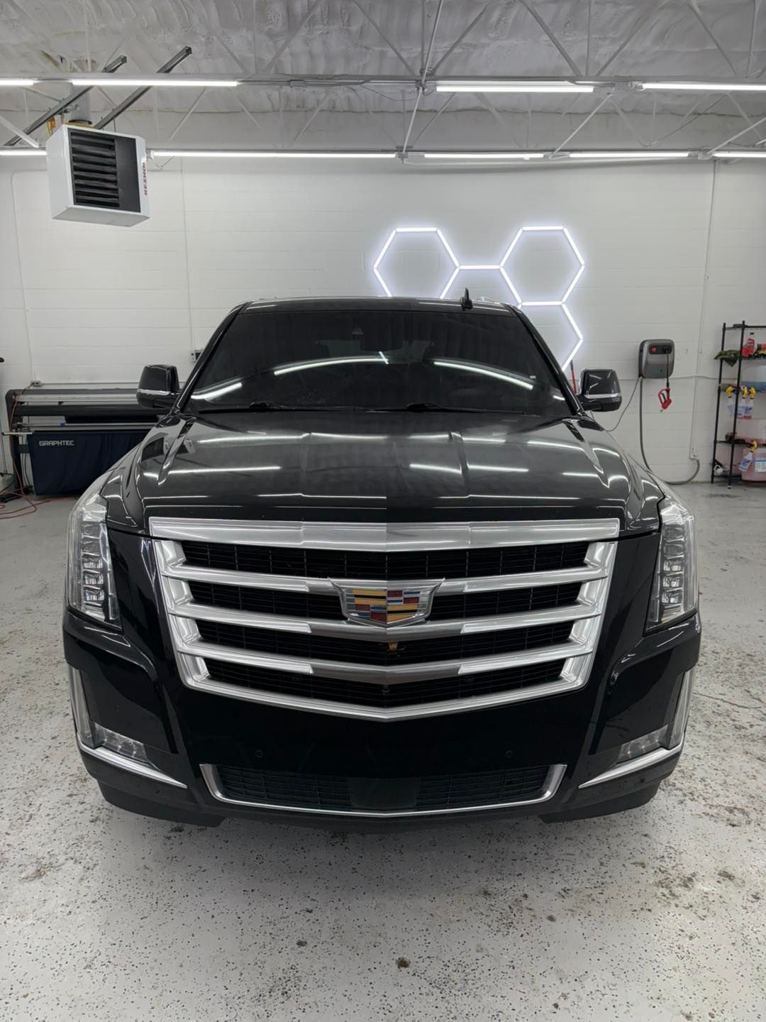 A black Cadillac Escalade SUV parked in a bright, white-walled garage with hexagon wall lights.