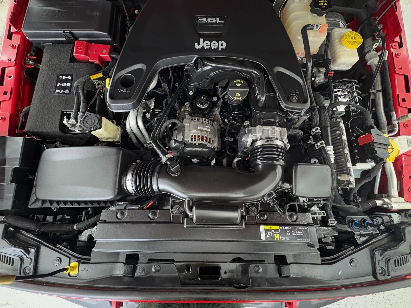 A high-angle view of the engine bay of a red Jeep, featuring a black engine cover with the Jeep logo and 3.6L label.