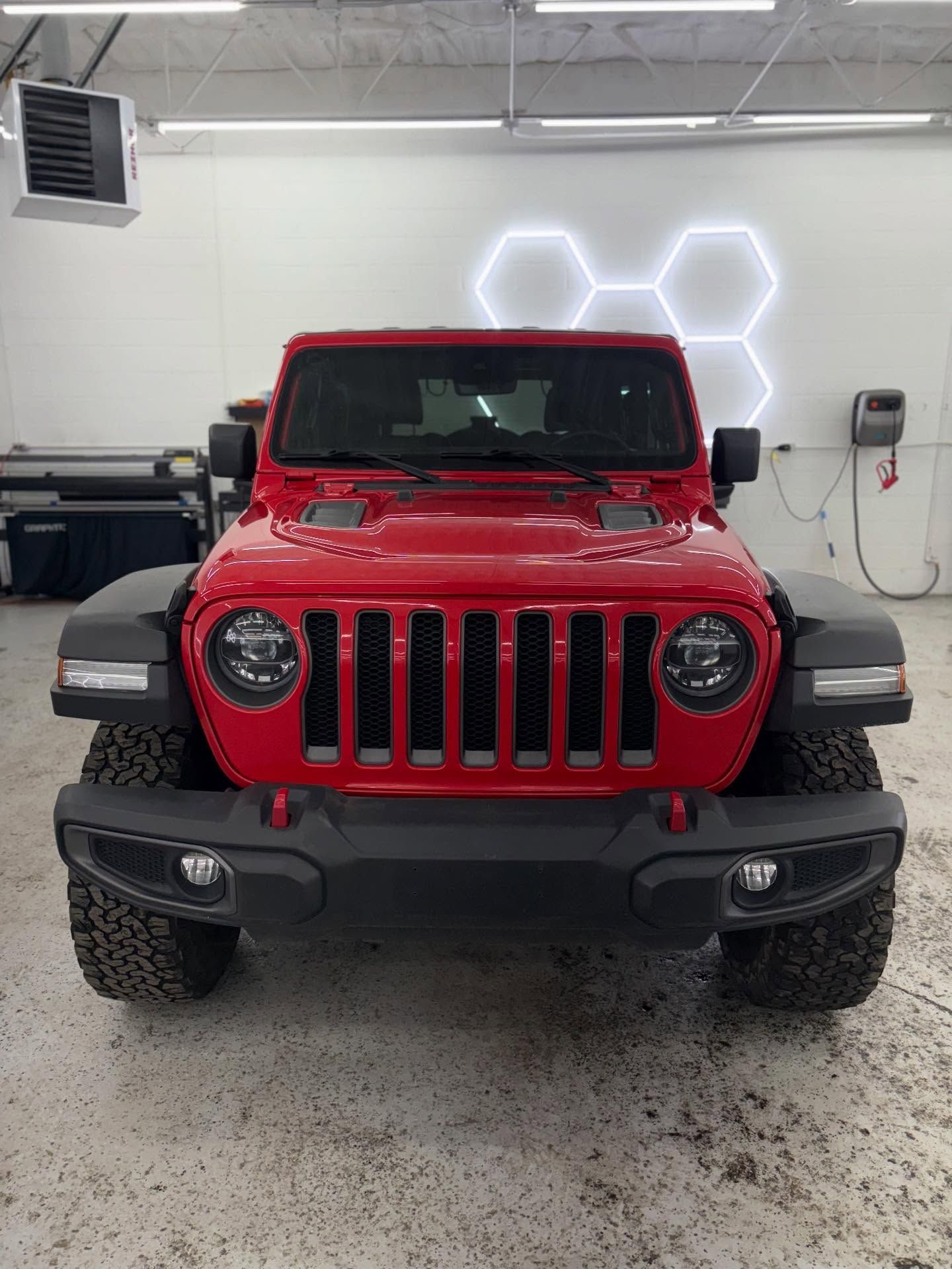 A bright red Jeep Wrangler parked facing forward in a clean, brightly lit indoor workshop with hexagon wall lighting.