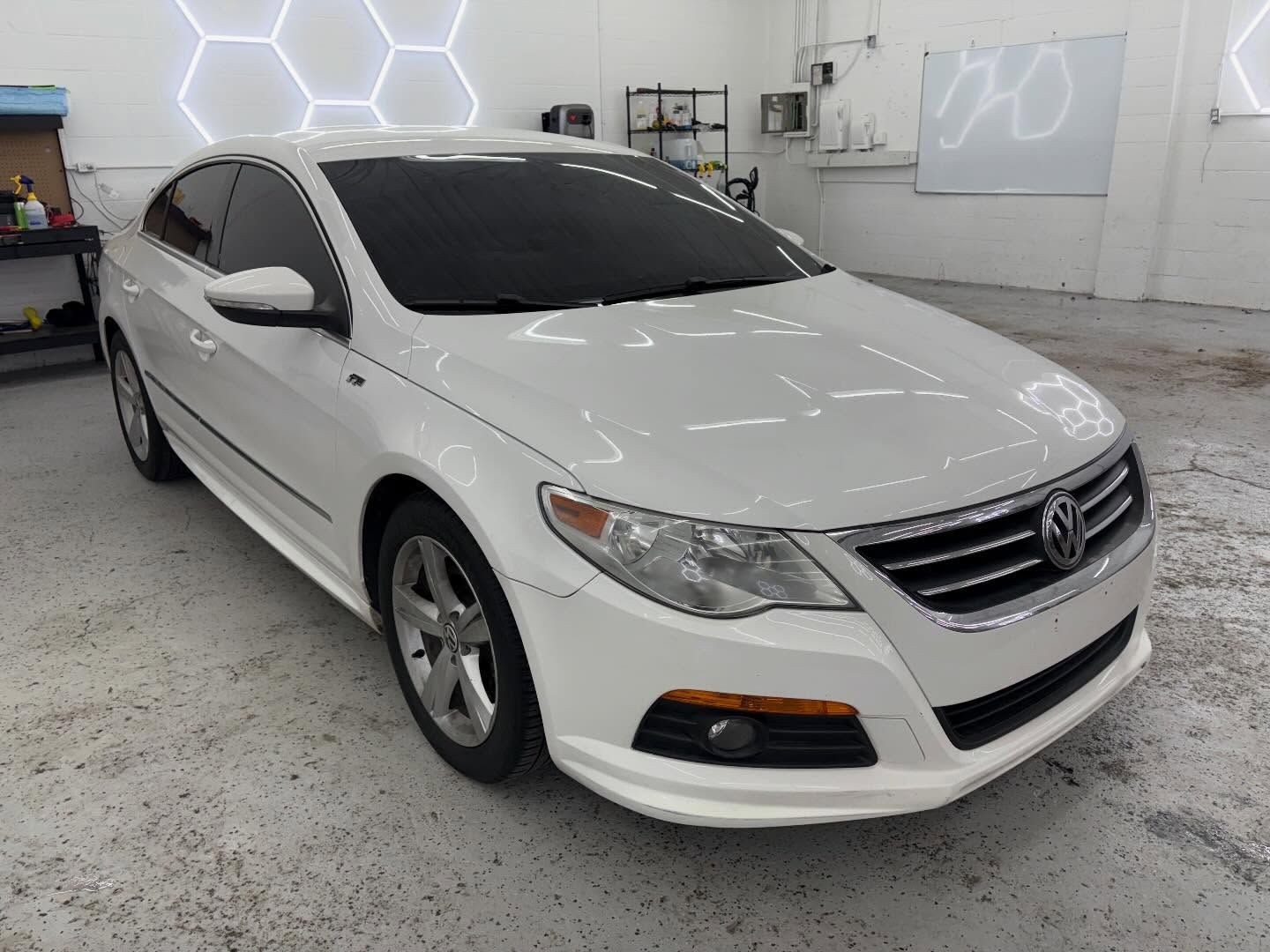 A white Volkswagen CC sedan parked at an angle inside a well-lit automotive workshop with hexagonal wall lighting.